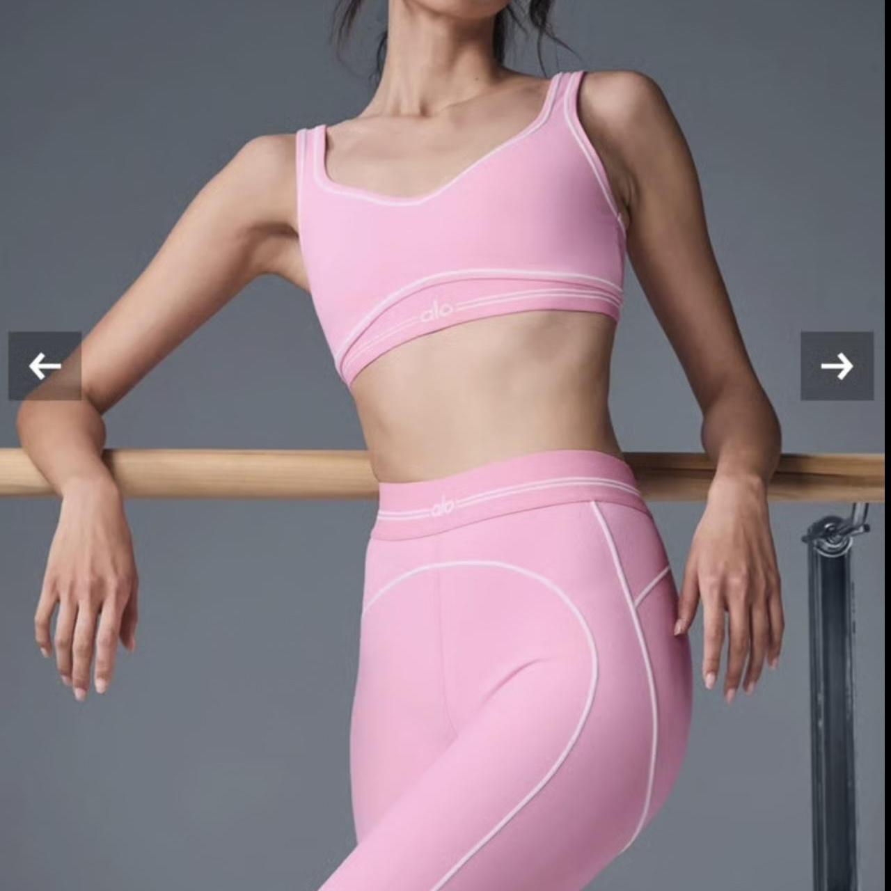 Woman wearing a matching light pink Alo Yoga sports bra and high-waisted leggings set, standing at a ballet barre with arms relaxed.