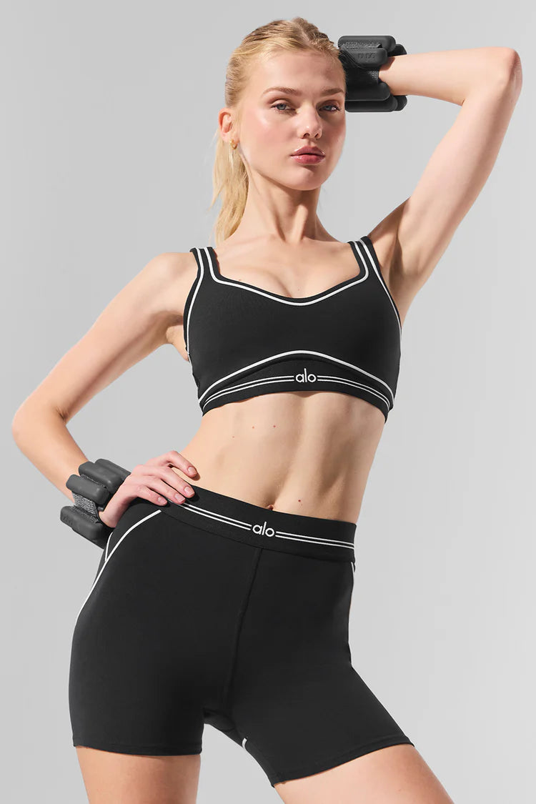 Woman posing in black Alo workout set with white trim, including sports bra and shorts, wearing black wrist weights against gray backdrop.