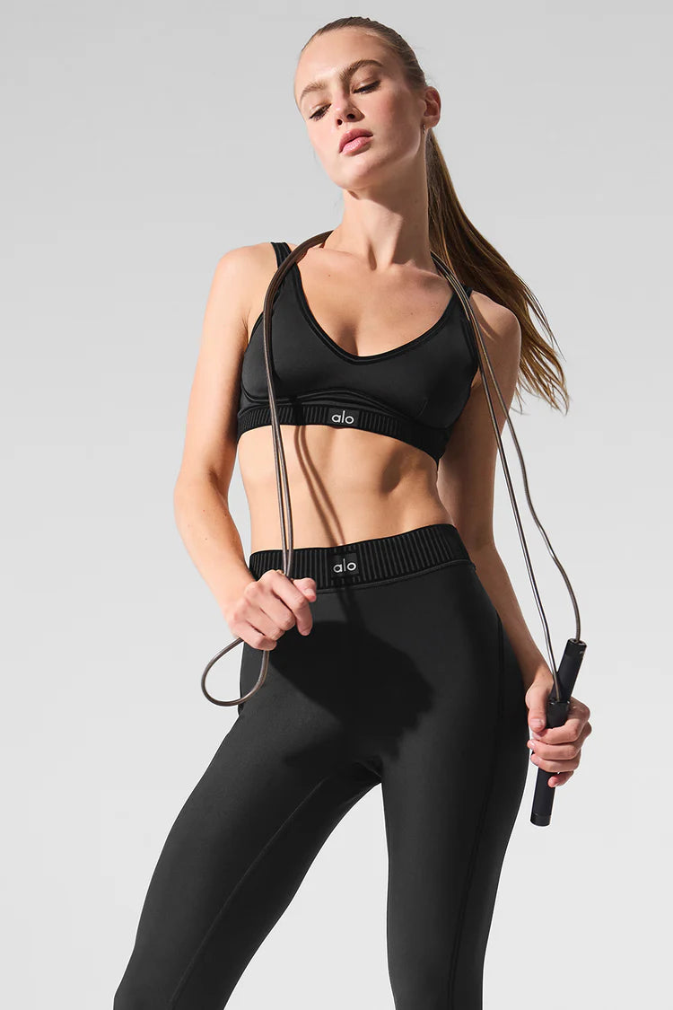 A woman wearing a black Alo sports bra and leggings, holding a jump rope while standing against a neutral background.