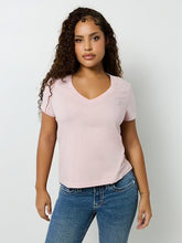 Woman with curly hair wearing a light pink V-neck T-shirt and blue jeans standing against a white background, facing forward.