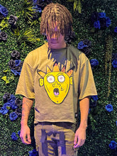 Young man with dreadlocks wearing an oversized olive green shirt with a cartoon face design, standing before a green floral wall.