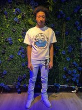 A person wearing a white t-shirt with a cartoon character face, light blue pants, and white sneakers, standing in front of a blue floral backdrop.
