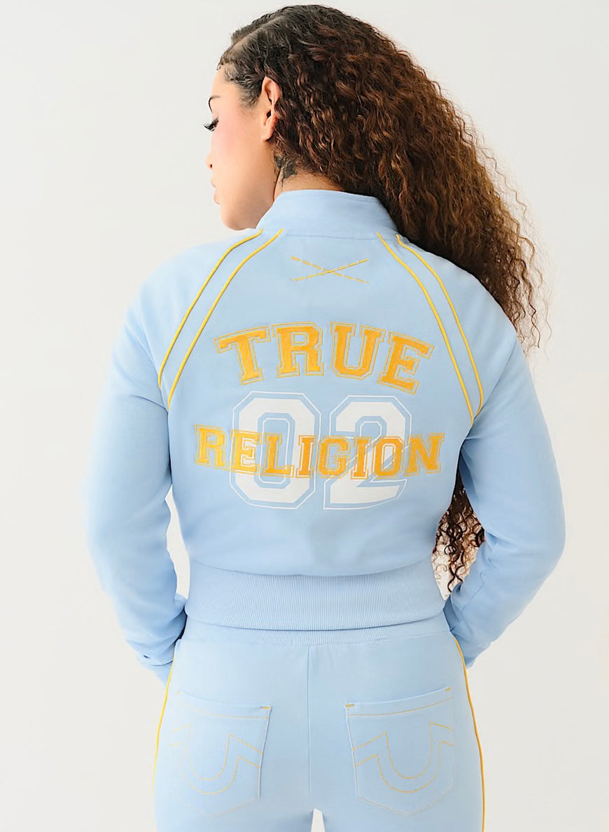 Woman wearing light blue zip-up track jacket with yellow piping and “TRUE RELIGION” text, paired with matching high-waisted joggers.