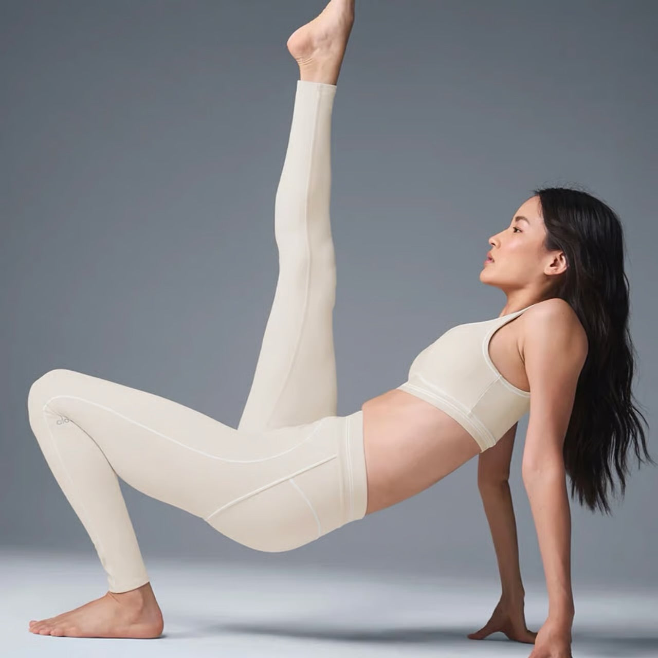 A woman in beige athletic wear performs a yoga pose, lifting one leg high while supporting her body with hands and feet.