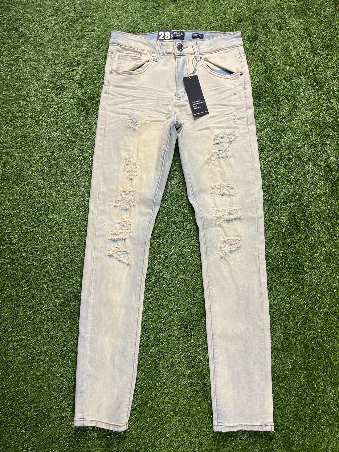 Light-wash distressed skinny jeans with ripped details on the front, displayed flat on a green grass background, waist size 28.