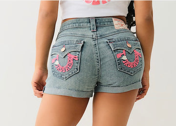 Back view of a person wearing light denim shorts with pink "True Religion" embroidery on pockets and a white cropped shirt.