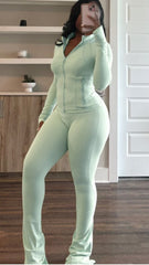 Woman wearing light mint green zip-up two-piece set with flared pants, taking mirror selfie in room with wooden floors and shelves.