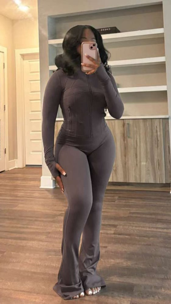 Woman with long curly hair wearing a fitted, long-sleeve, dark gray jumpsuit standing barefoot on wooden floor indoors.