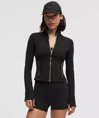 Woman wearing black zip-up athletic jacket with gold zippers, matching black shorts, and black cap, posing against plain light background.