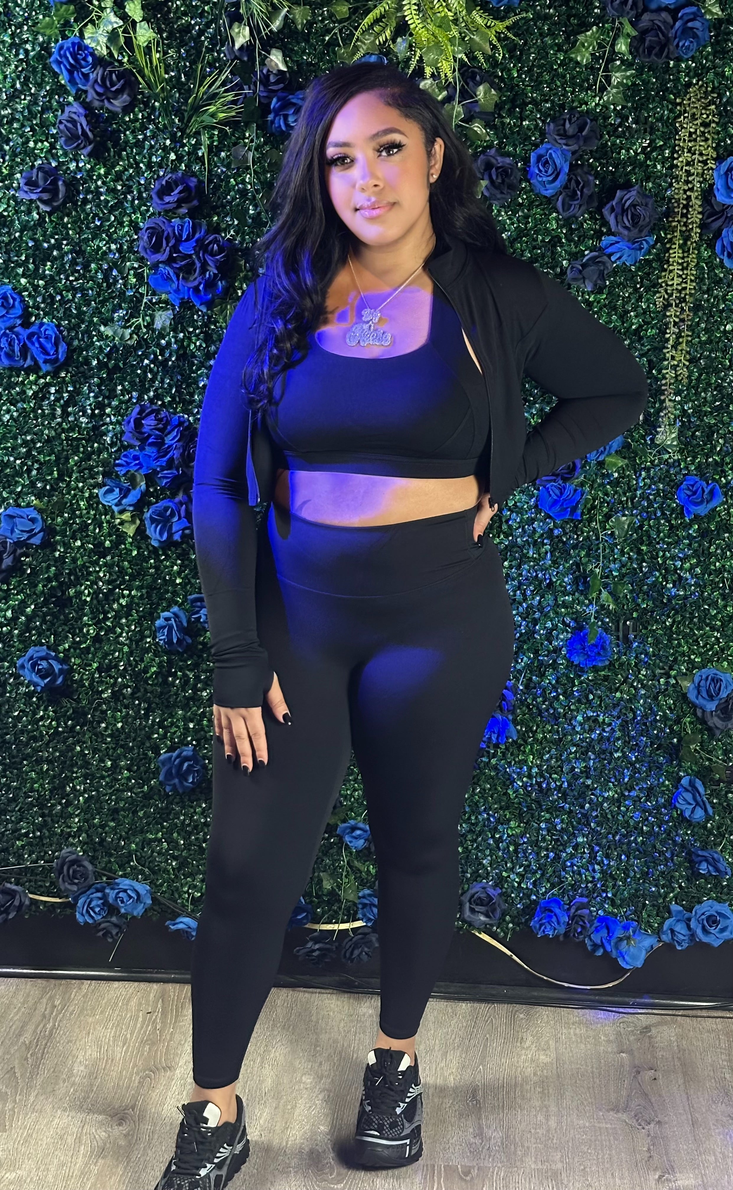 A woman in a black crop top, jacket, leggings, and sneakers stands against a green wall decorated with blue and black flowers.