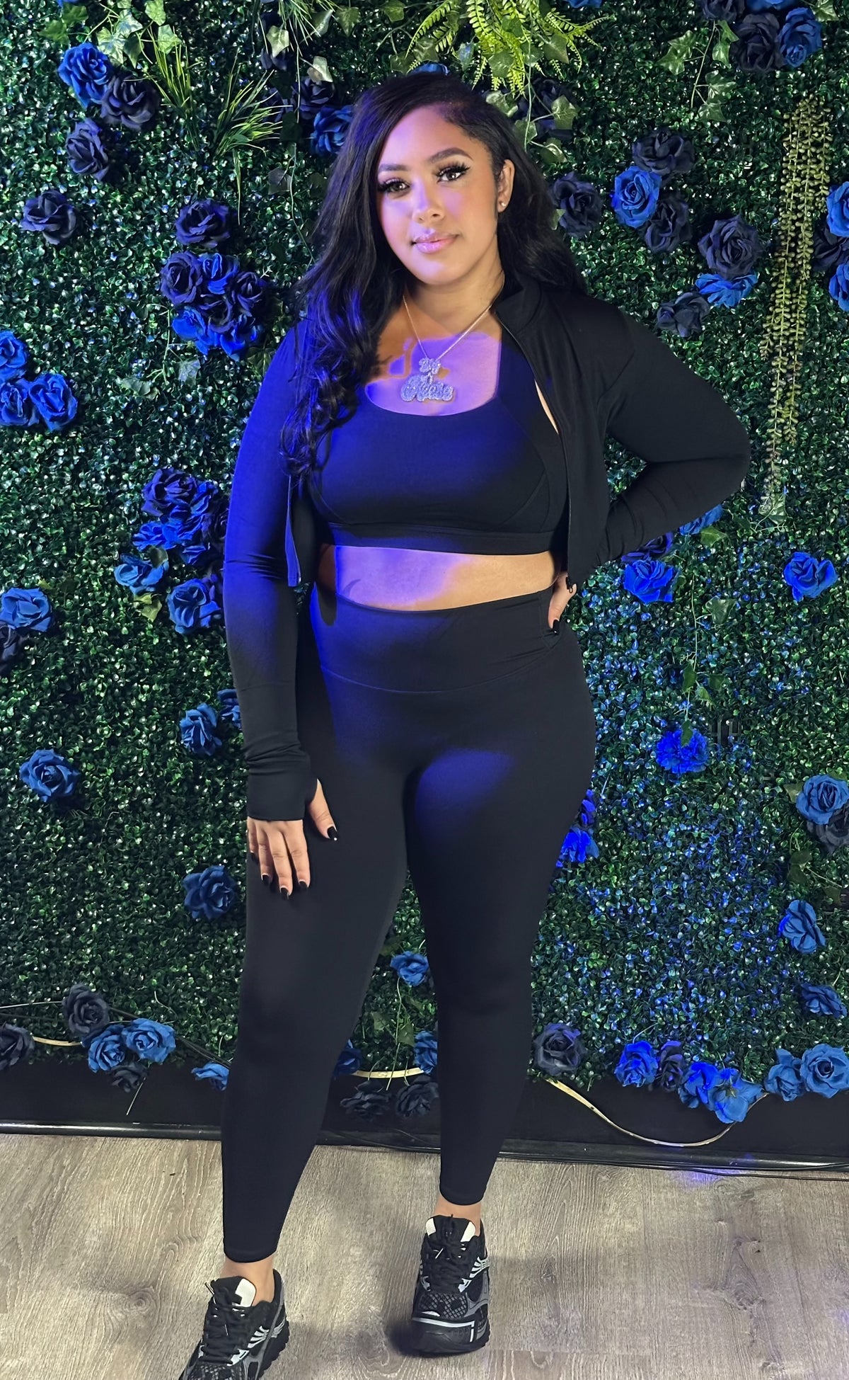 A woman in a black crop top, jacket, leggings, and sneakers stands against a green wall decorated with blue and black flowers.