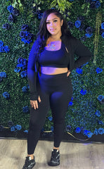 A woman in a black crop top, jacket, leggings, and sneakers stands against a green wall decorated with blue and black flowers.