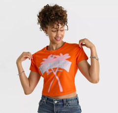 Smiling woman wearing an orange crop top with white palm tree graphic and “True Religion Brand Jeans” text, paired with denim jeans.