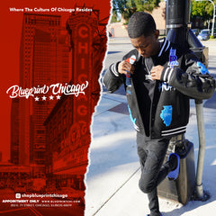 Man wearing a black varsity jacket with colorful patches, black ripped jeans, and a chain, standing on a city street with a Chicago backdrop.