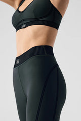 A close-up of a person wearing a dark green Alo sports bra and matching leggings with a ribbed waistband, showing the side view.