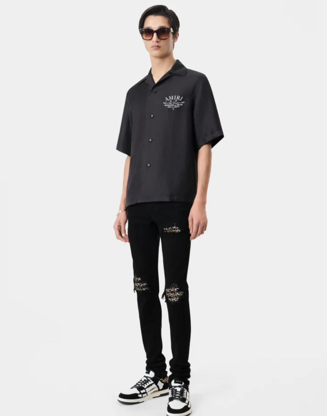 A man wearing an AMIRI short-sleeve shirt, ripped black jeans, and black and white sneakers, paired with sunglasses.