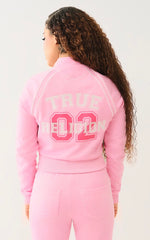 Woman wearing a matching pink tracksuit with "TRUE RELIGION 02" printed on the back of the jacket, shown from behind with long curly hair.