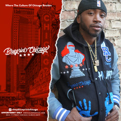 A man wearing a black hoodie with a multicolored "Blueprint Chicago" varsity jacket, standing against a brick wall.