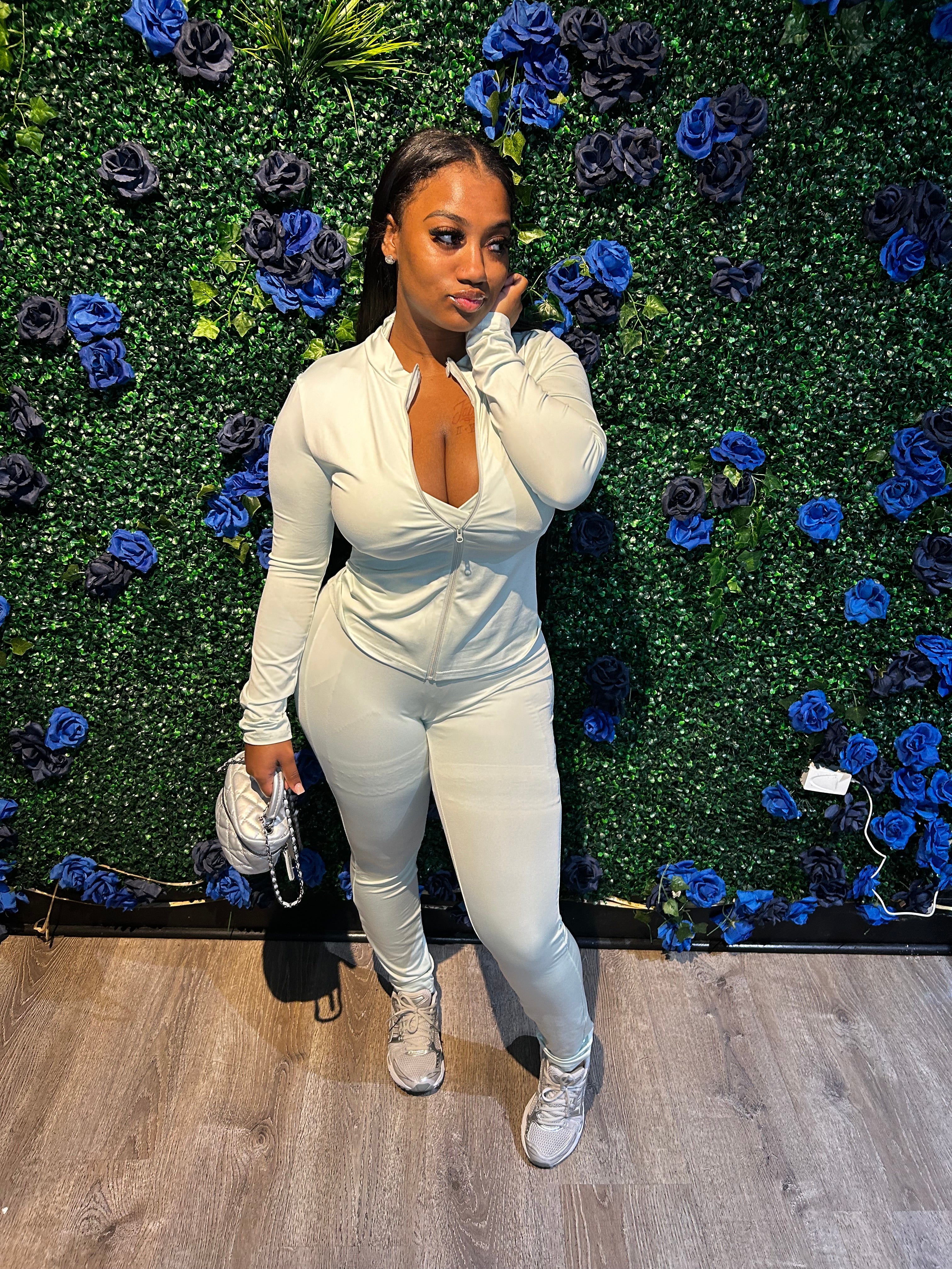 A woman in a light beige zip-up bodysuit, paired with sneakers, holding a small handbag, standing in front of a floral backdrop.