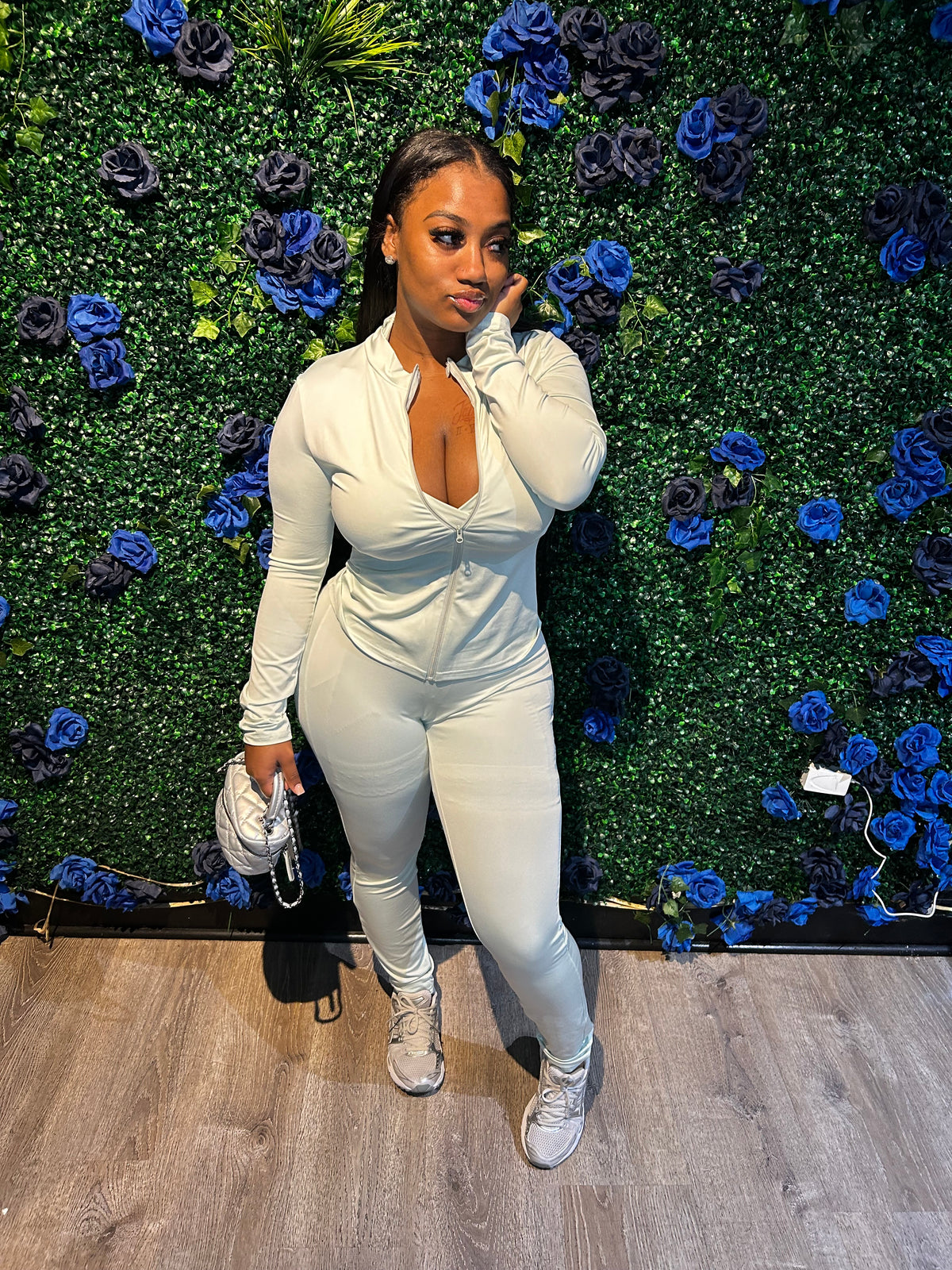 A woman in a light beige zip-up bodysuit, paired with sneakers, holding a small handbag, standing in front of a floral backdrop.