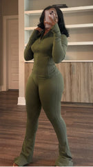 Woman in green zip-up jacket and matching pants taking mirror selfie in room with wooden floor and shelves in the background.