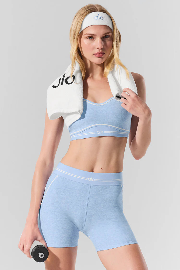 Woman in light blue Alo workout set with matching shorts and sports bra, wearing headband, holding towel and dumbbell, posing confidently.