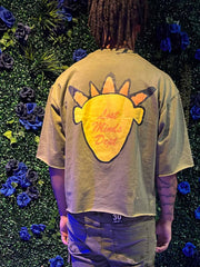 Person wearing light brown cropped t-shirt with yellow and orange design and text “Lost Minds Dept,” standing by blue flowers.