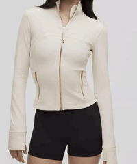 Woman wearing a fitted cream zip-up jacket with gold zipper details, paired with black shorts, standing against a plain background.