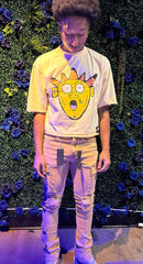 A person wearing a yellow t-shirt with a cartoon face design, paired with beige cargo pants, and black tags hanging from the pants, standing in front of a floral backdrop.