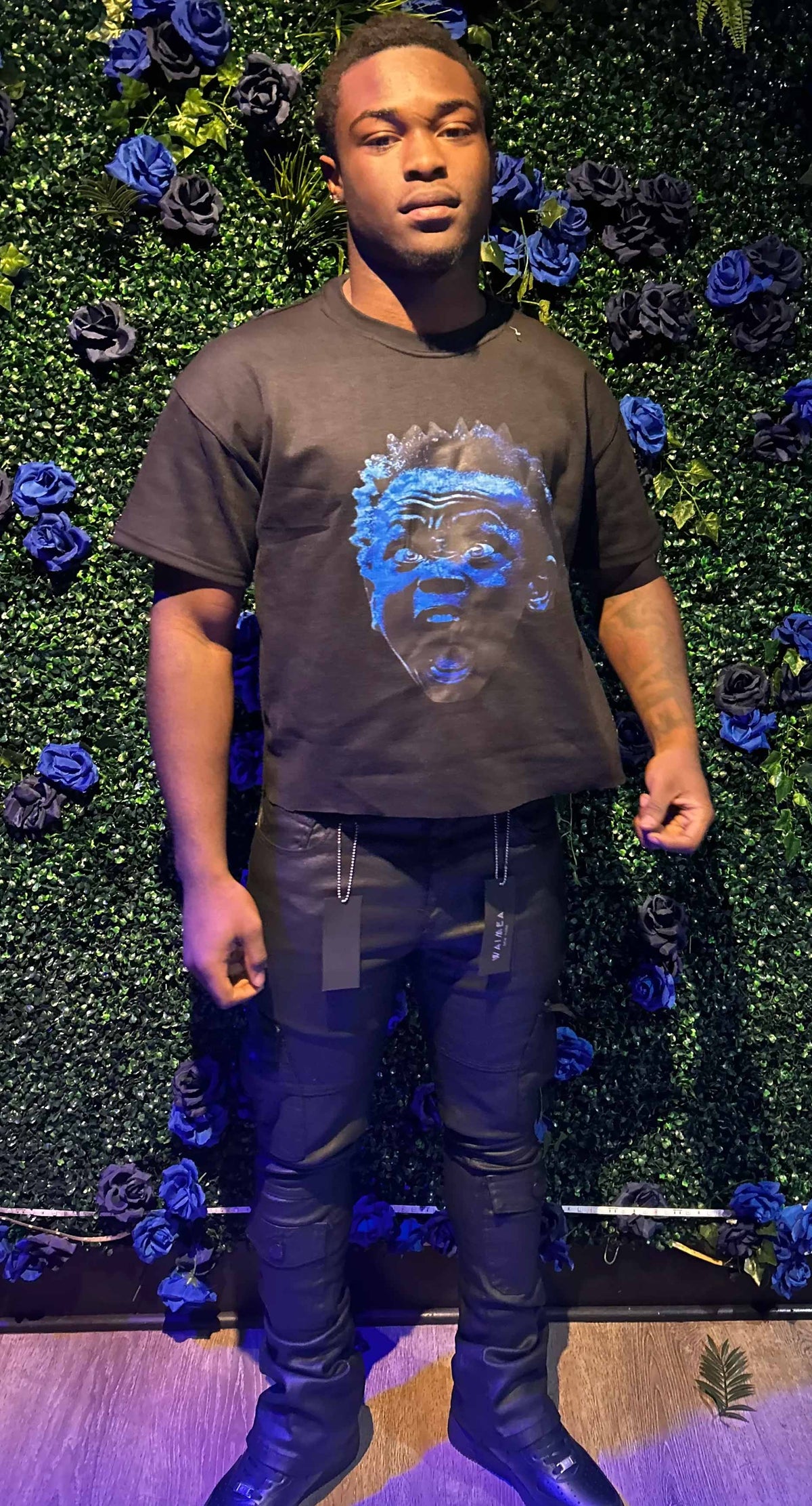 Man stands in front of green wall with blue and black roses, wearing black shirt with blue face print and black pants.