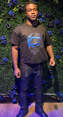 Man stands in front of green wall with blue and black roses, wearing black shirt with blue face print and black pants.