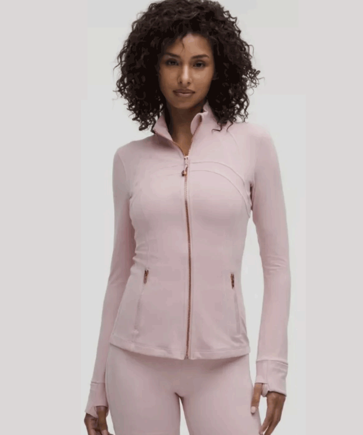 Woman with curly hair wearing light pink zip-up jacket and matching pants, posing against plain light gray background.