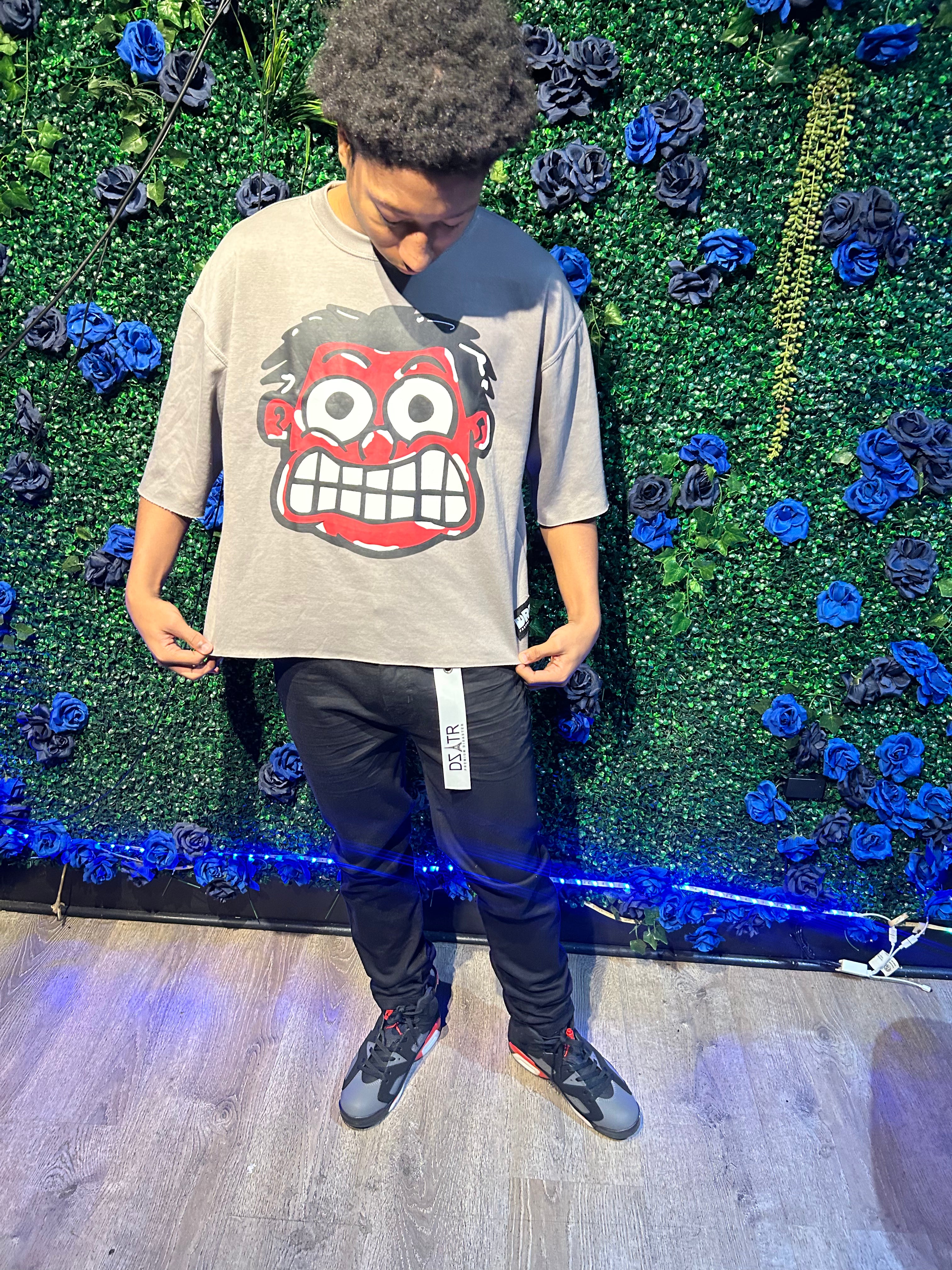 Man wearing a gray t-shirt with a large cartoon face, black pants with a tag, and black and gray sneakers, standing by flowers.