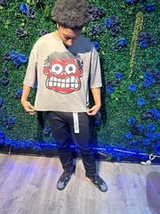 Man wearing a gray t-shirt with a large cartoon face, black pants with a tag, and black and gray sneakers, standing by flowers.