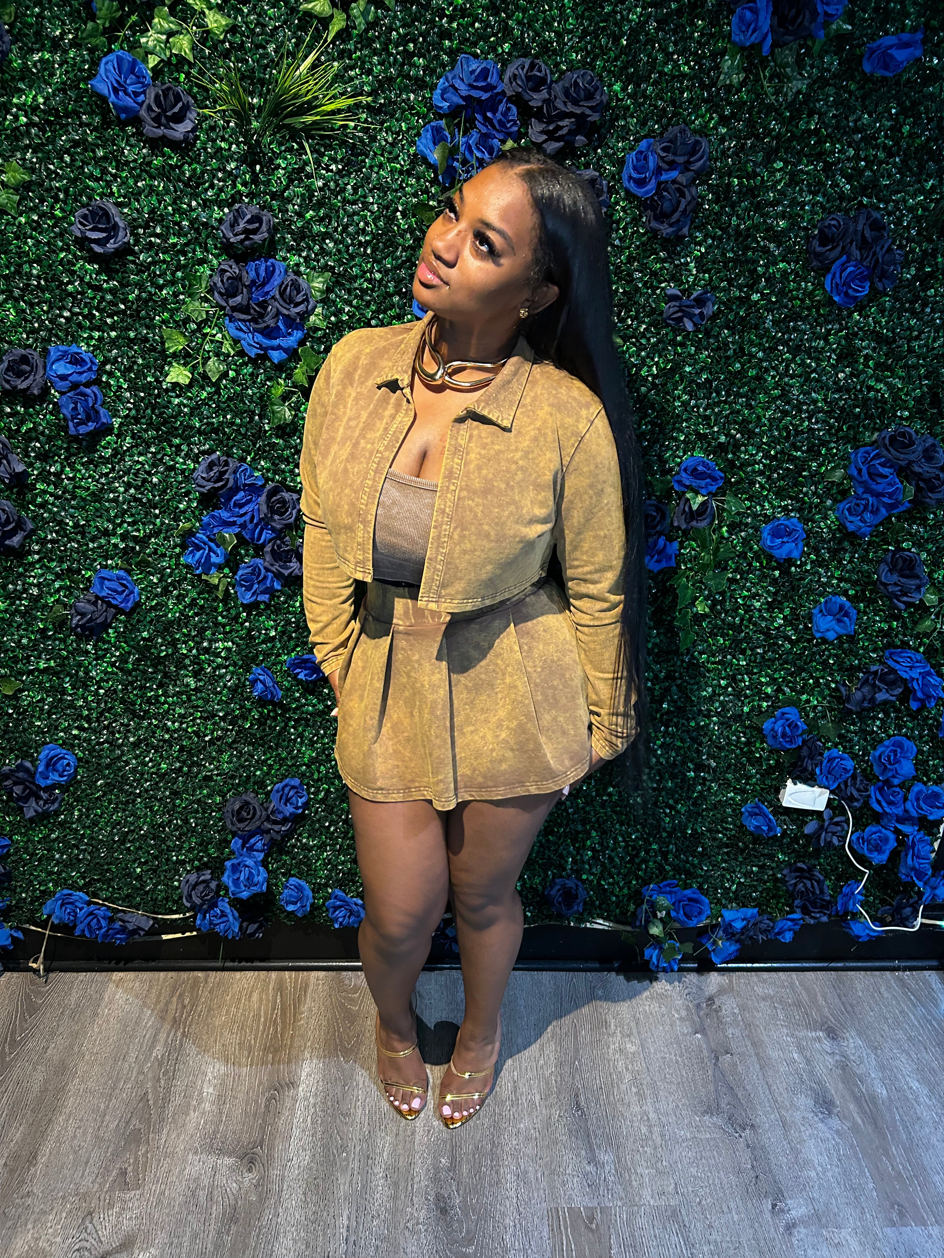 Woman in tan outfit with gold heels poses on wooden floor against green floral wall with blue and black roses, looking slightly upward.