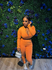 Woman in an orange zip-up two-piece outfit holding a silver quilted handbag, posing in front of a green floral wall with blue roses.