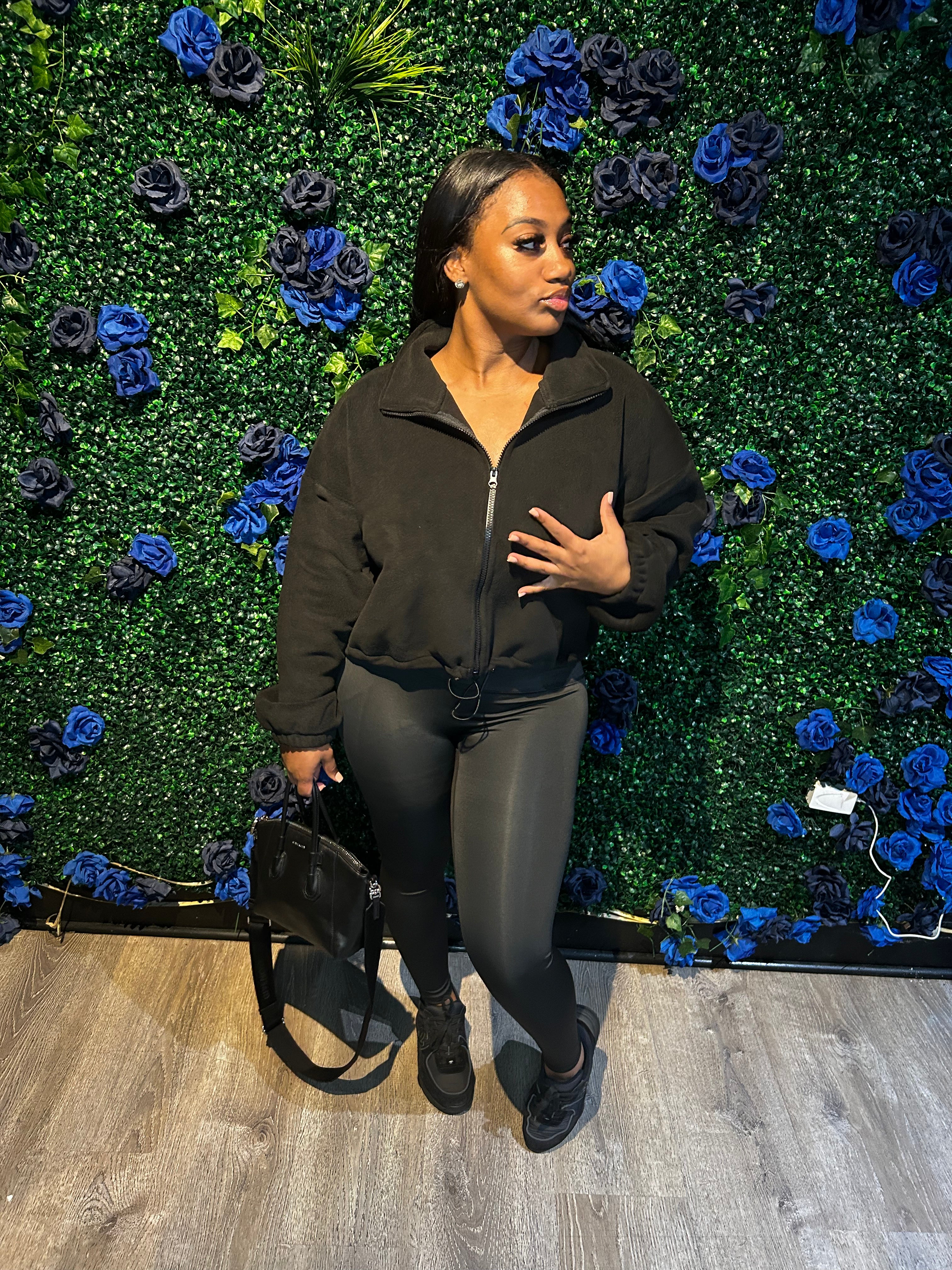 Woman wearing a black zip-up jacket, black leggings, black sneakers, and holding a black bag, standing in front of a floral green wall.
