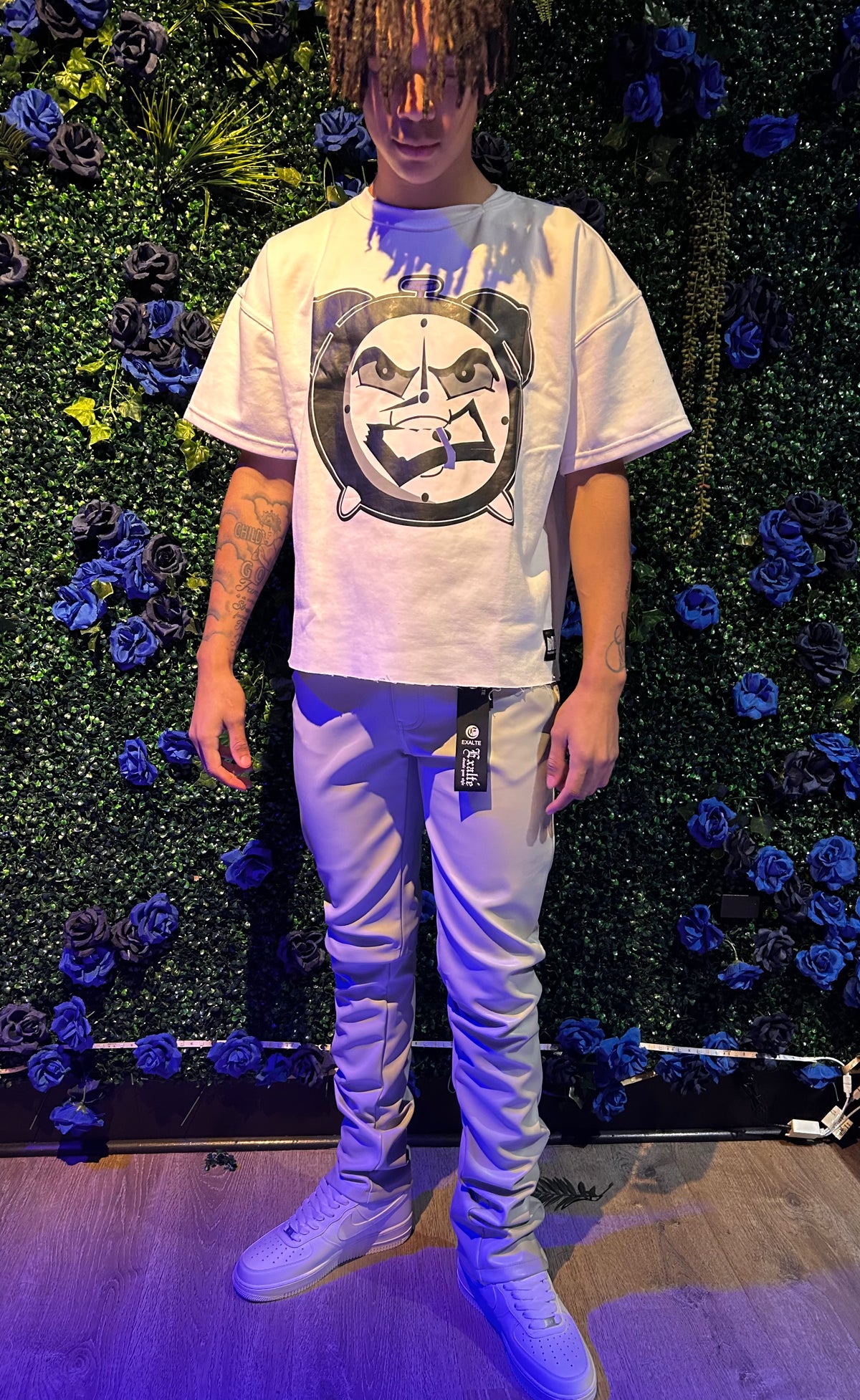 Man wearing oversized white graphic t-shirt with angry clock print, slim-fit beige pants, and white sneakers, standing by floral green wall.