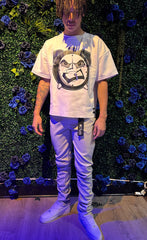 Man wearing oversized white graphic t-shirt with angry clock print, slim-fit beige pants, and white sneakers, standing by floral green wall.