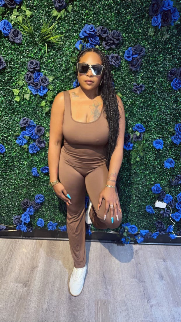 Woman poses in a brown sleeveless jumpsuit and white sneakers against a green floral wall with blue and black roses, wearing sunglasses.