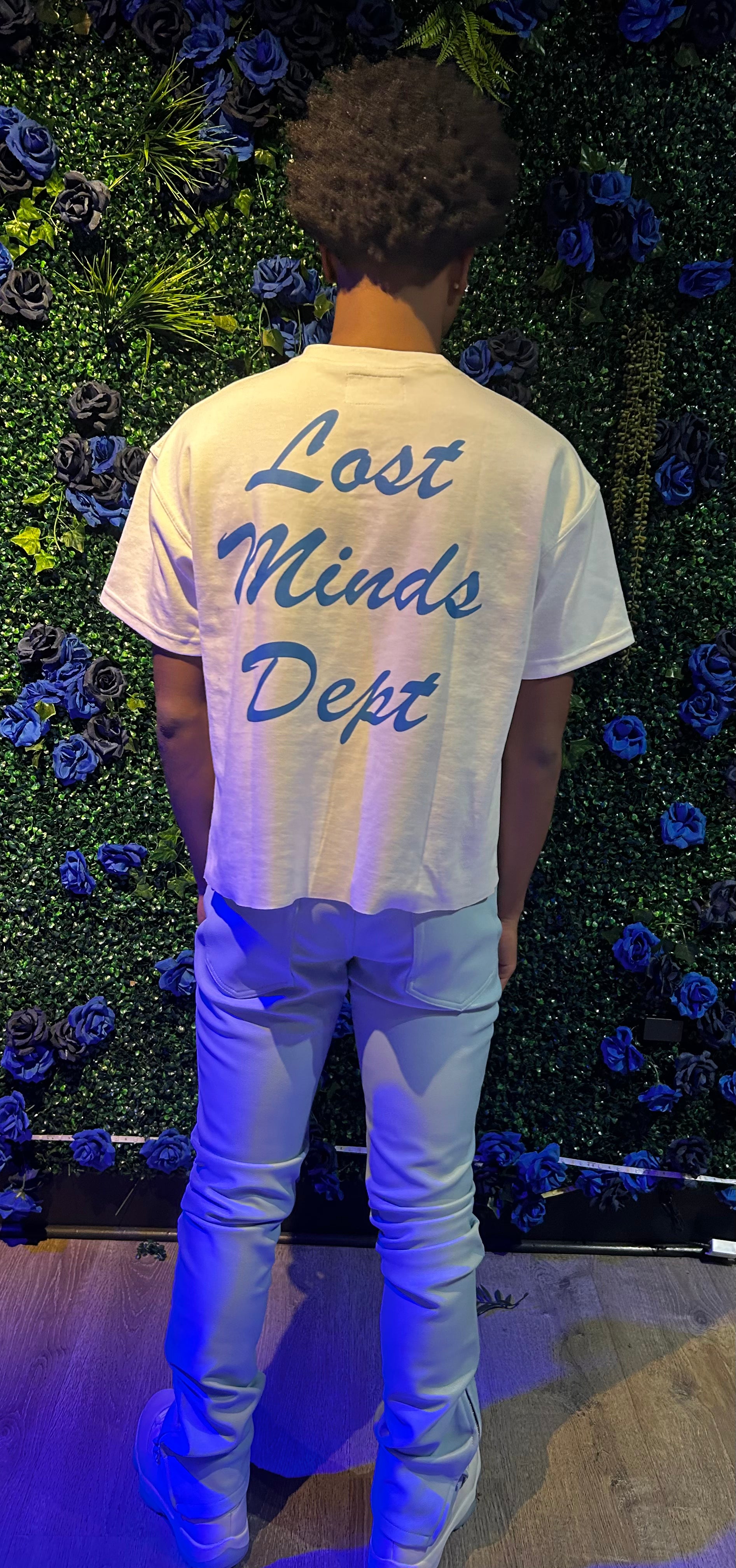 Person standing with back to camera wearing white shirt with “Lost Minds Dept” in blue, white pants, and white sneakers.