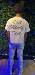 Person standing with back to camera wearing white shirt with “Lost Minds Dept” in blue, white pants, and white sneakers.