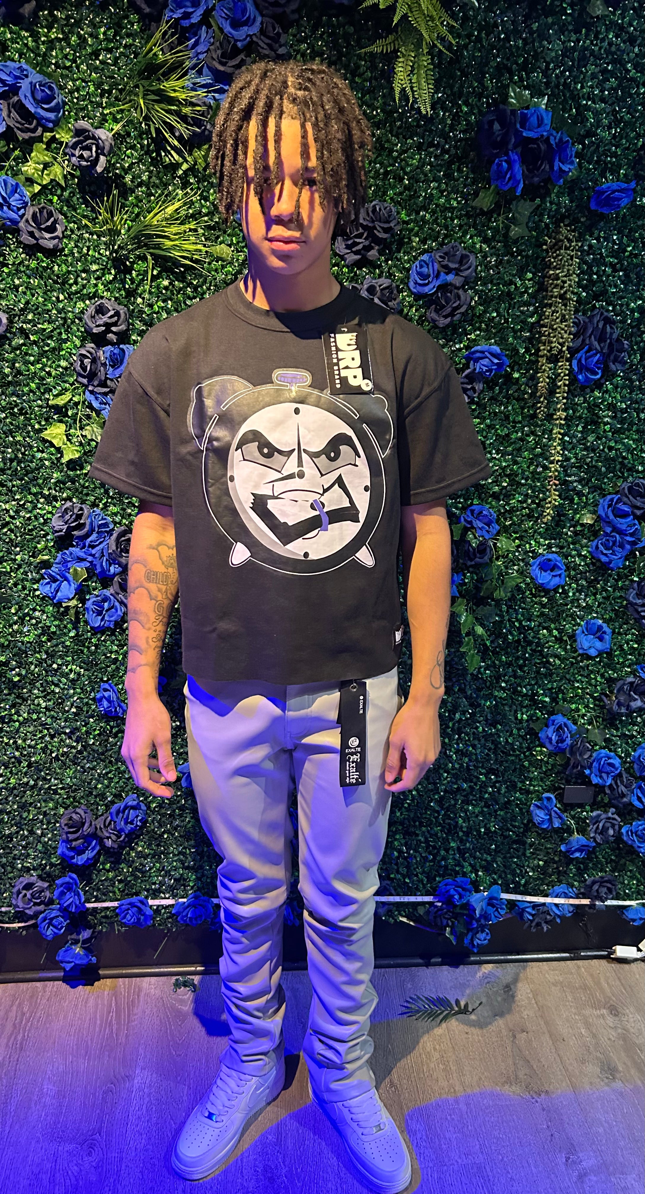 A person wearing a black graphic t-shirt with a clock design, beige pants, and white sneakers, standing in front of a blue floral backdrop.