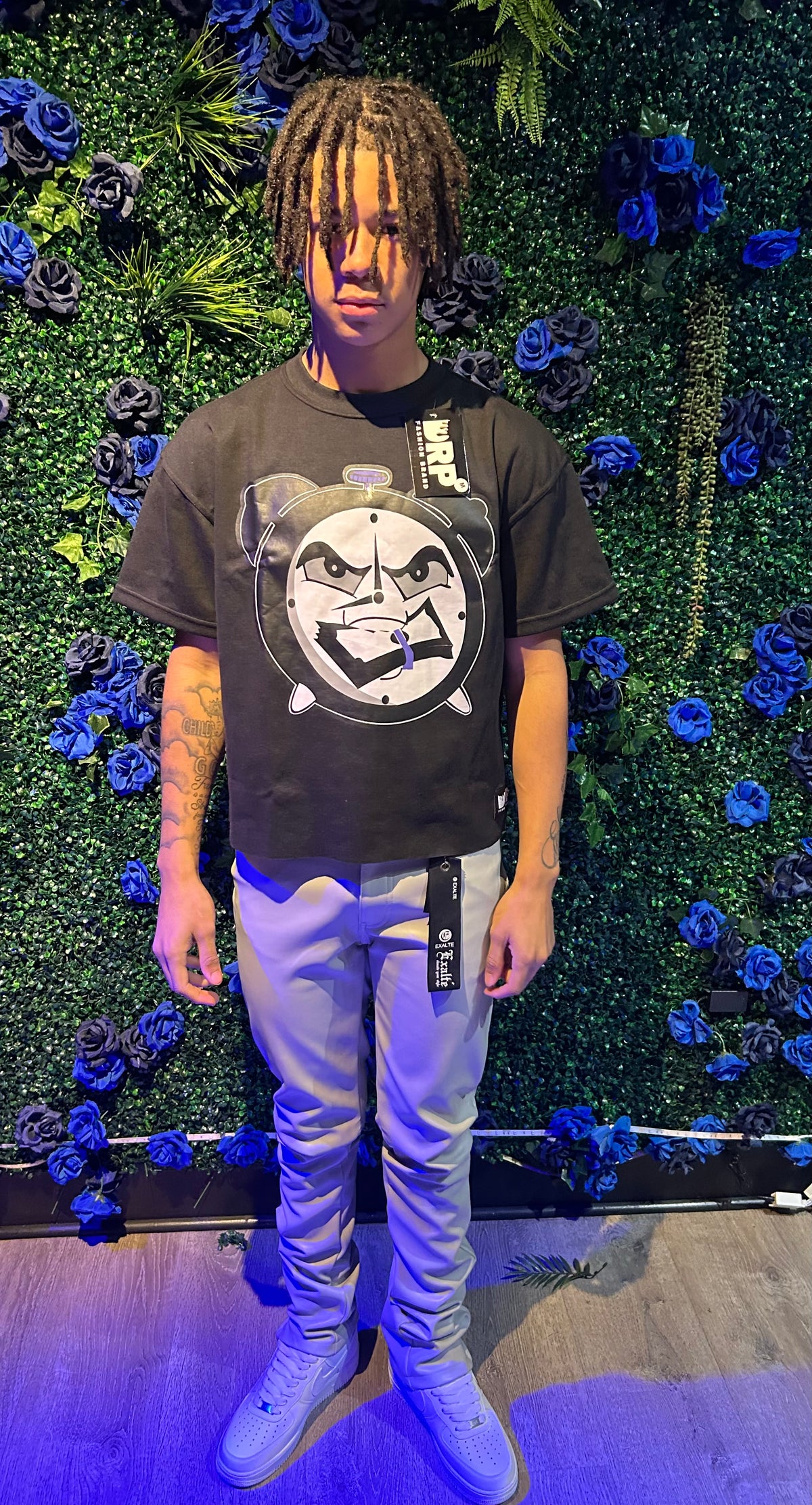 A person wearing a black graphic t-shirt with a clock design, beige pants, and white sneakers, standing in front of a blue floral backdrop.