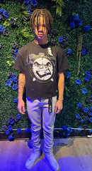 A person wearing a black graphic t-shirt with a clock design, beige pants, and white sneakers, standing in front of a blue floral backdrop.